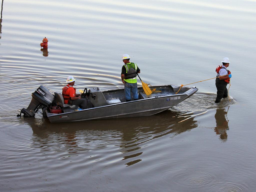 Three workers in safety gear stand with a small motorboat in shallow water; one’s paddling, another’s steering, and one walks while pulling the boat with a rope.