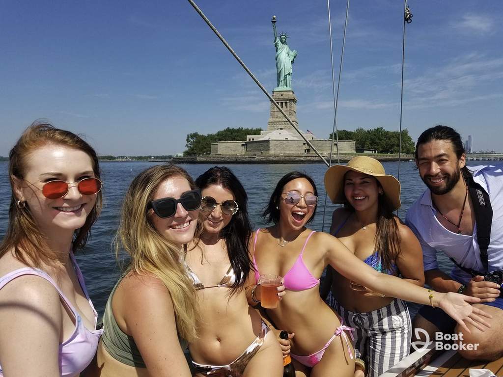 Six people in swimwear smile on a boat in front of the Statue of Liberty on a sunny day, with water and blue sky behind them.