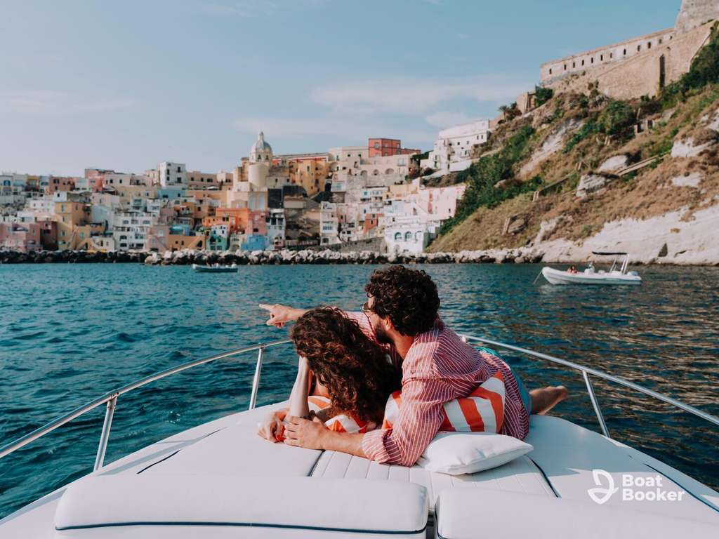 A couple’s relaxing at the bow of a boat, gazing toward a vibrant coastal town built on a hillside under a clear sky.
