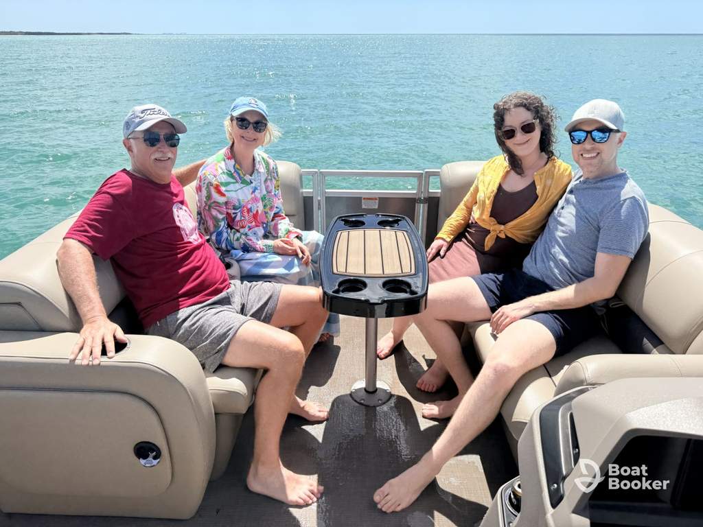 Four people are sitting on a boat in sunny weather, smiling at the camera. The sea’s calm and blue, with a small table between them. A “Boat Booker” logo’s in the bottom right corner.