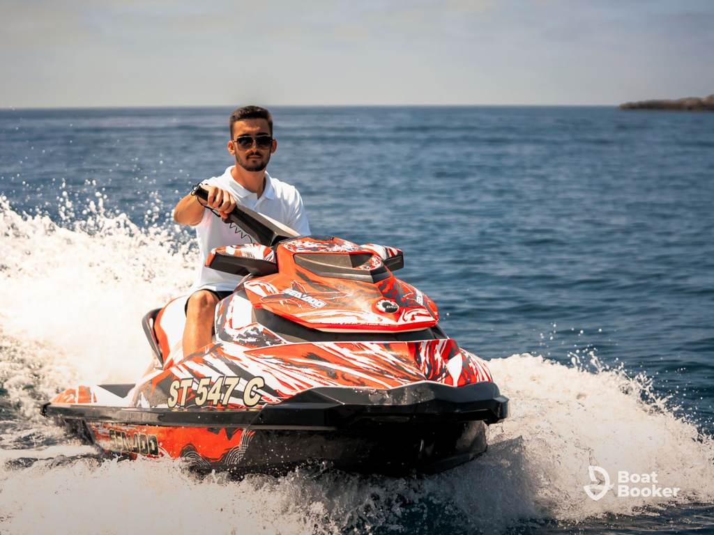 A man’s riding a red and white jet ski across the ocean on a sunny day, creating a spray of water behind him.