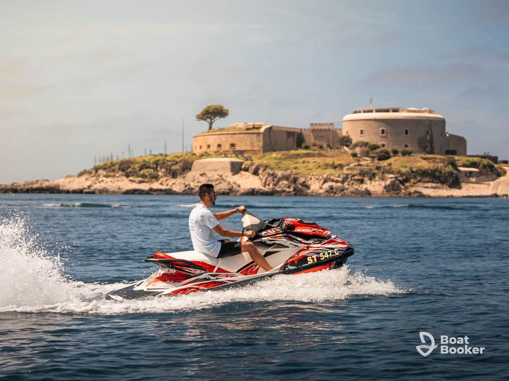 Someone’s riding a red jet ski on calm water near a small island with a historic stone fortress, all under a clear sky.