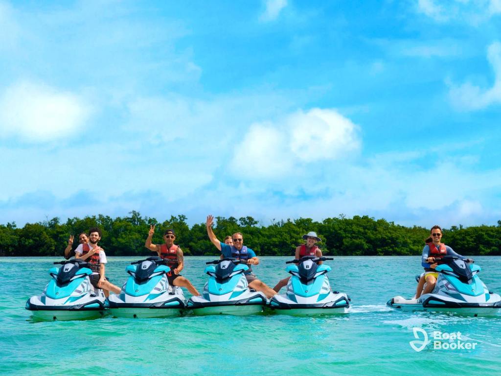Five people on jet skis sit in a row on clear water, waving at the camera, with green trees and a blue sky in the background.