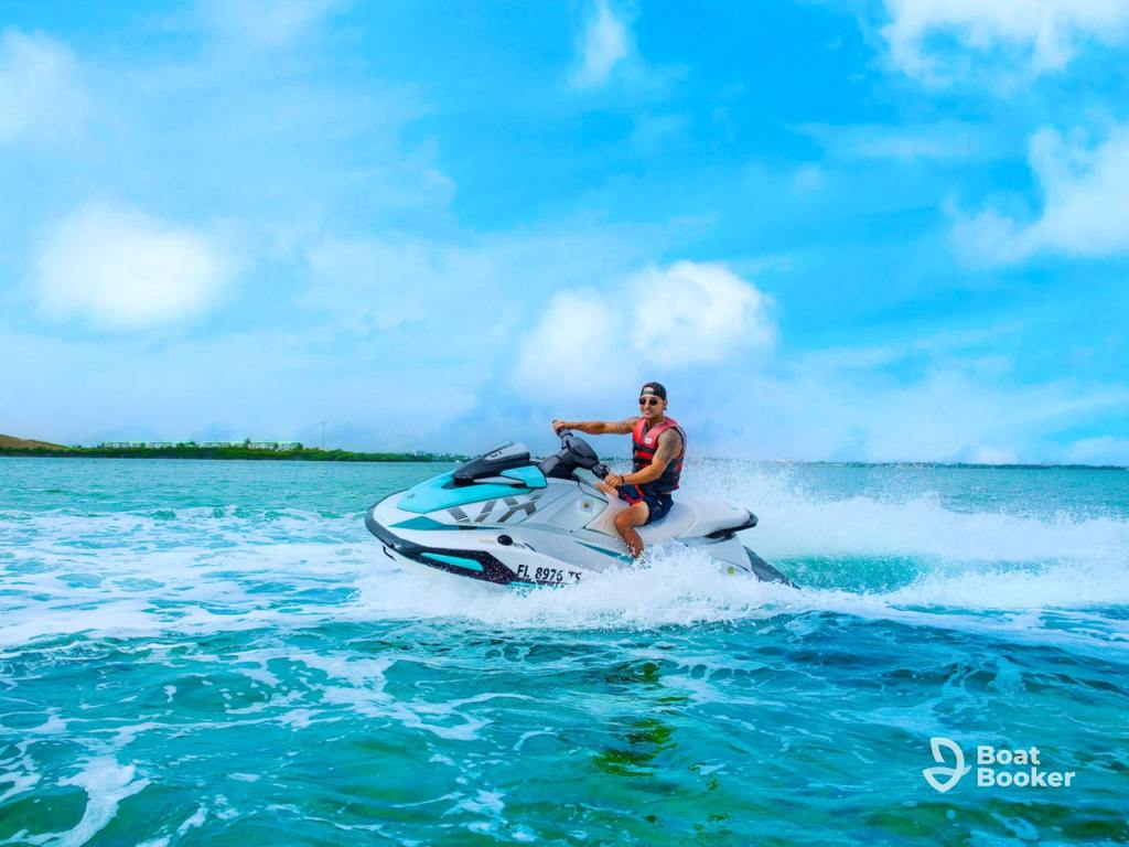 Someone’s riding a jet ski on blue-green water under a bright sky, making a white spray trail. The Boat Booker logo’s in the corner. No fish or species like Trout or Salmon are visible.