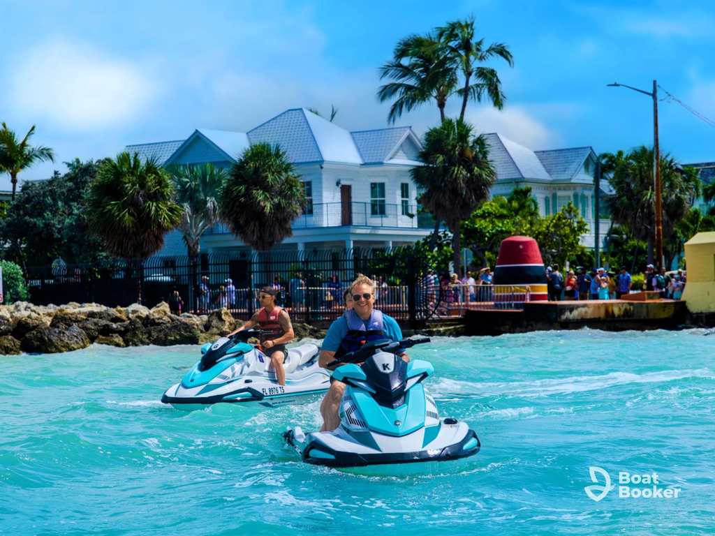 Two people ride jet skis on turquoise water by a shore with Palm Trees, a large white house, and a crowd in the background. The Boat Booker logo’s also visible.