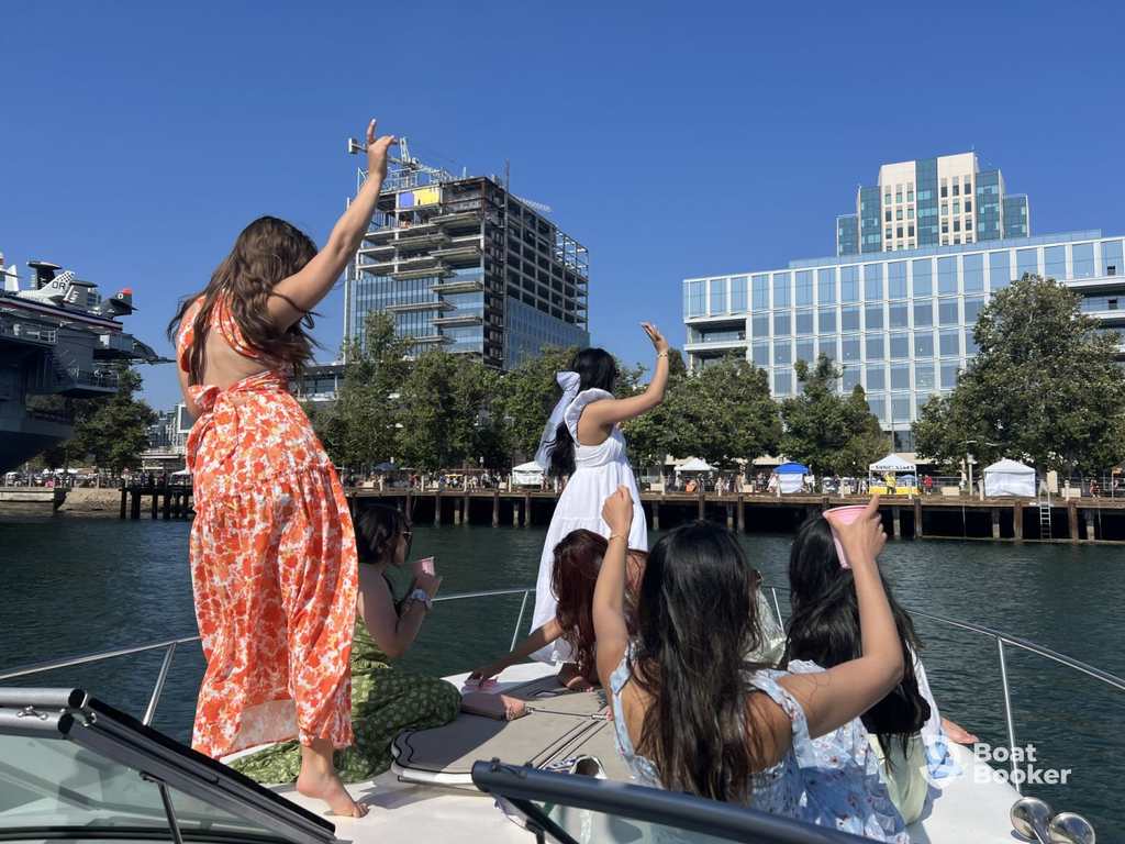 Five women on a boat raise their arms and wave toward the shore, with modern buildings and trees behind them under a clear blue sky.