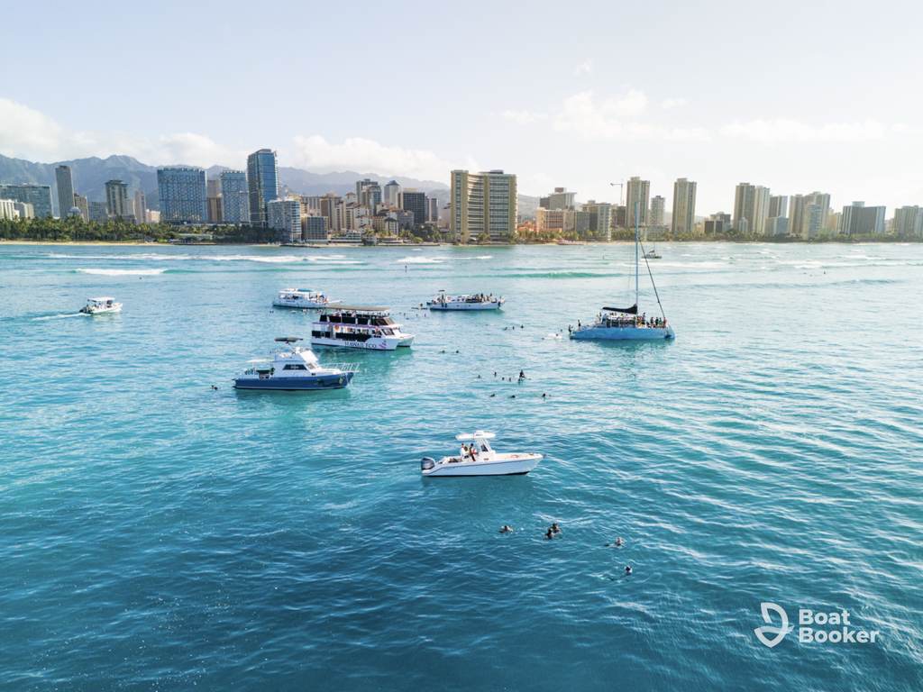 Several boats are anchored on clear blue water near a city skyline—perfect for island hopping in Hawaii. Swimmers enjoy the sea, and the “Boat Booker” logo’s in the lower right corner.