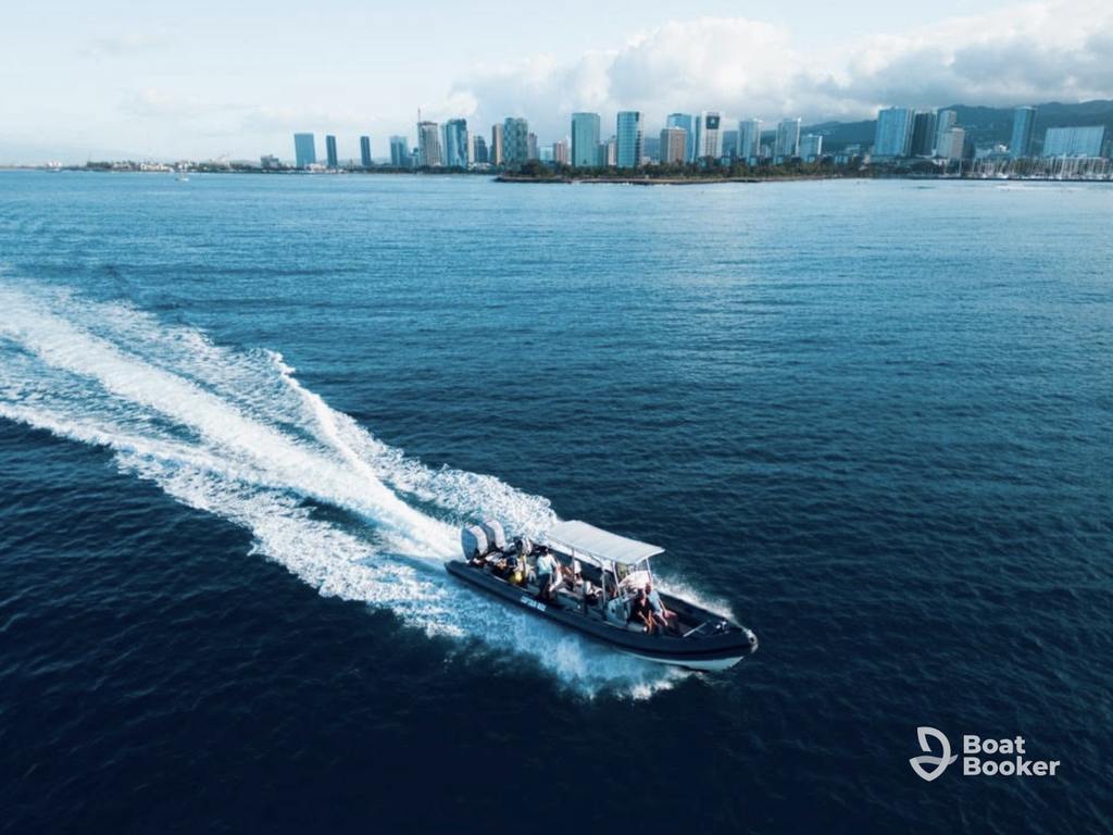 A motorboat with several passengers speeds across the water, leaving a wake—it’s perfect for island hopping in Hawaii, with a city skyline and mountains rising in the background.