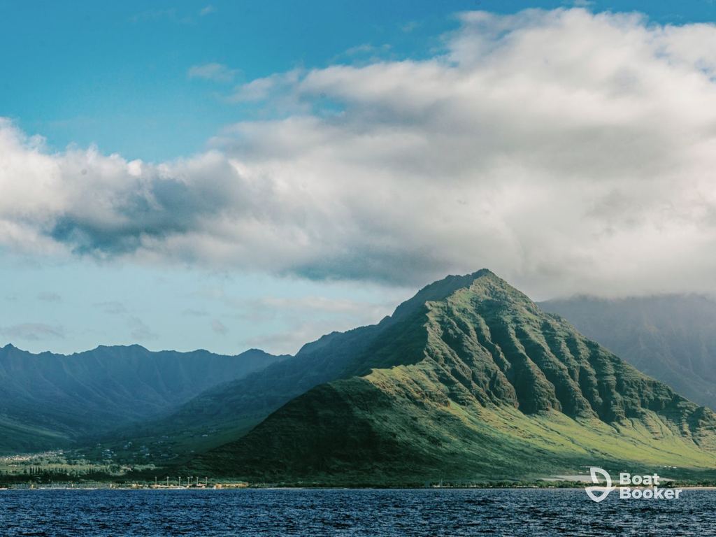 You’ll see a mountain with green slopes under a partly cloudy sky across water—ideal for island hopping in Hawaii. The “Boat Booker” logo’s in the bottom right corner.