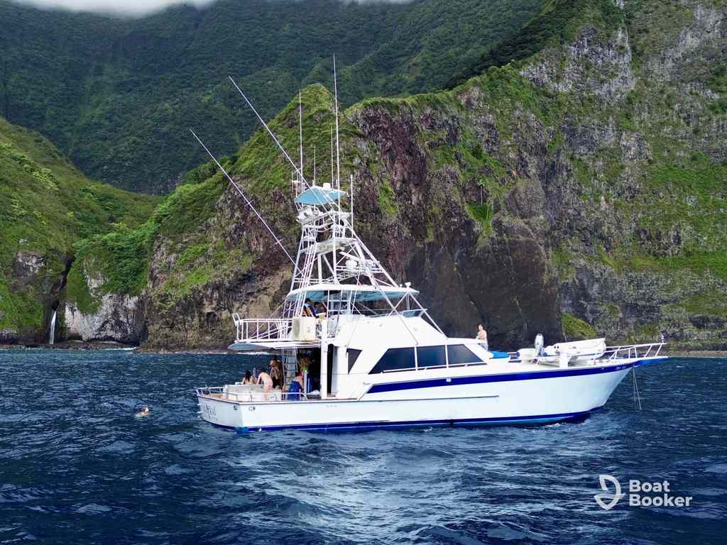 A white fishing boat with “Boat Booker” logo floats near a green coast—ideal for island hopping in Hawaii. There are several people on board, and one person’s swimming nearby.