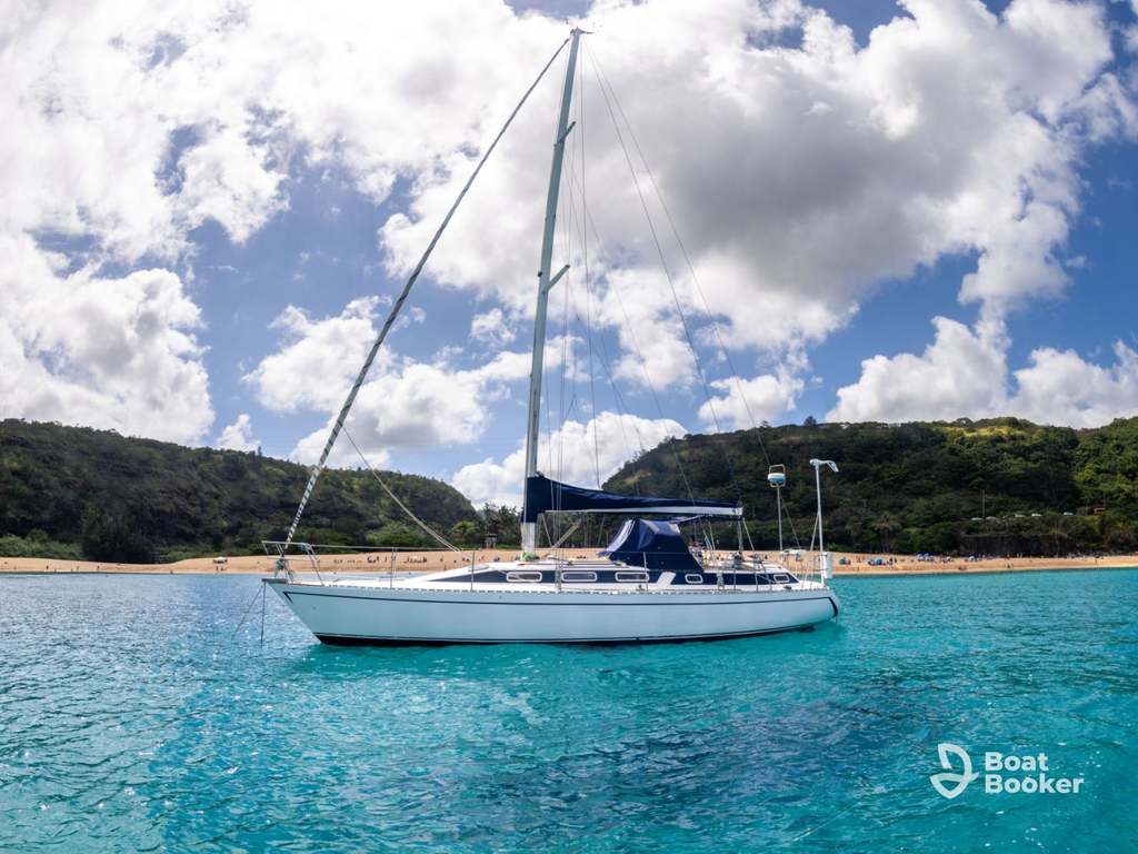 There’s a white sailboat anchored in clear turquoise water, ideal for island hopping in Hawaii, near a sandy beach with green hills and a partly cloudy sky behind.
