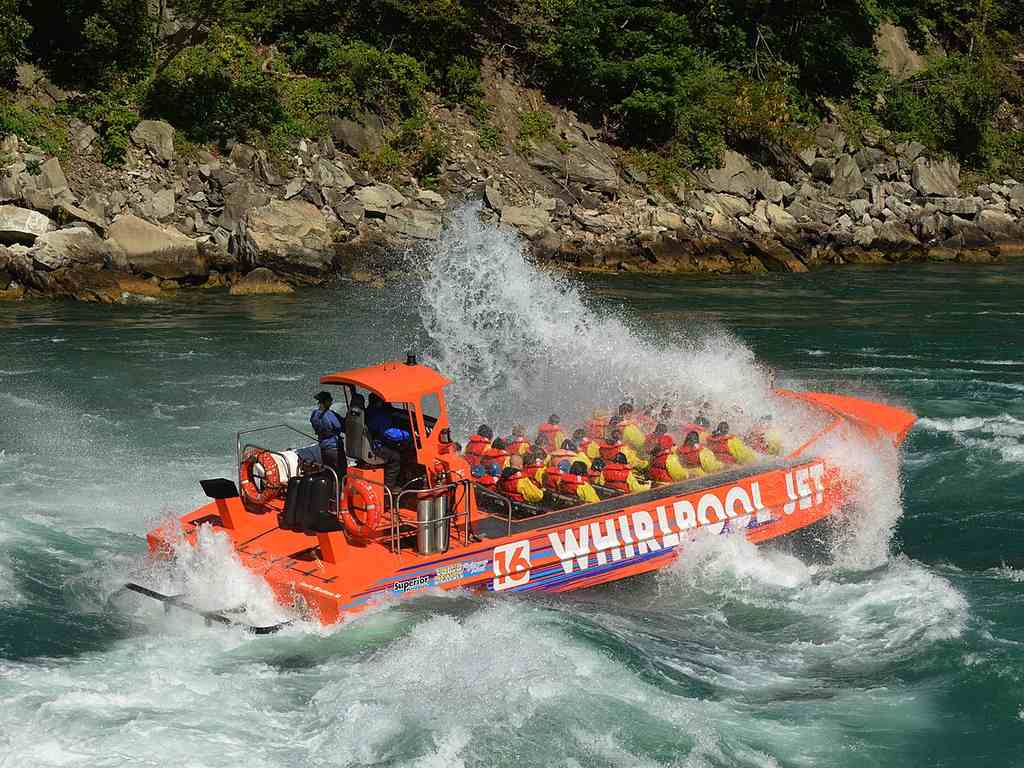 A bright orange jet boat with passengers in life jackets speeds through choppy river rapids, splashing water high by a rocky, tree-lined shore.