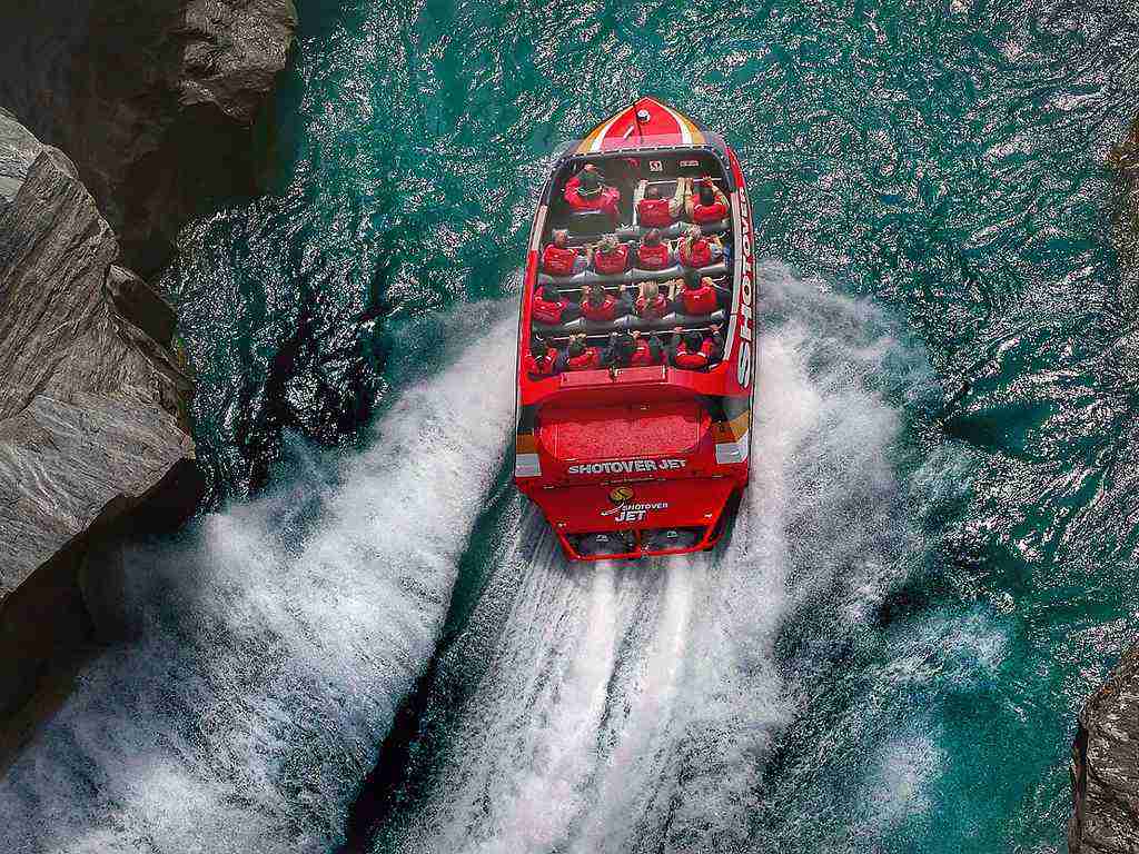 A red jet boat speeds through blue water between rocky cliffs, leaving a wake. Passengers wearing life jackets sit in rows—they’re enjoying the ride.