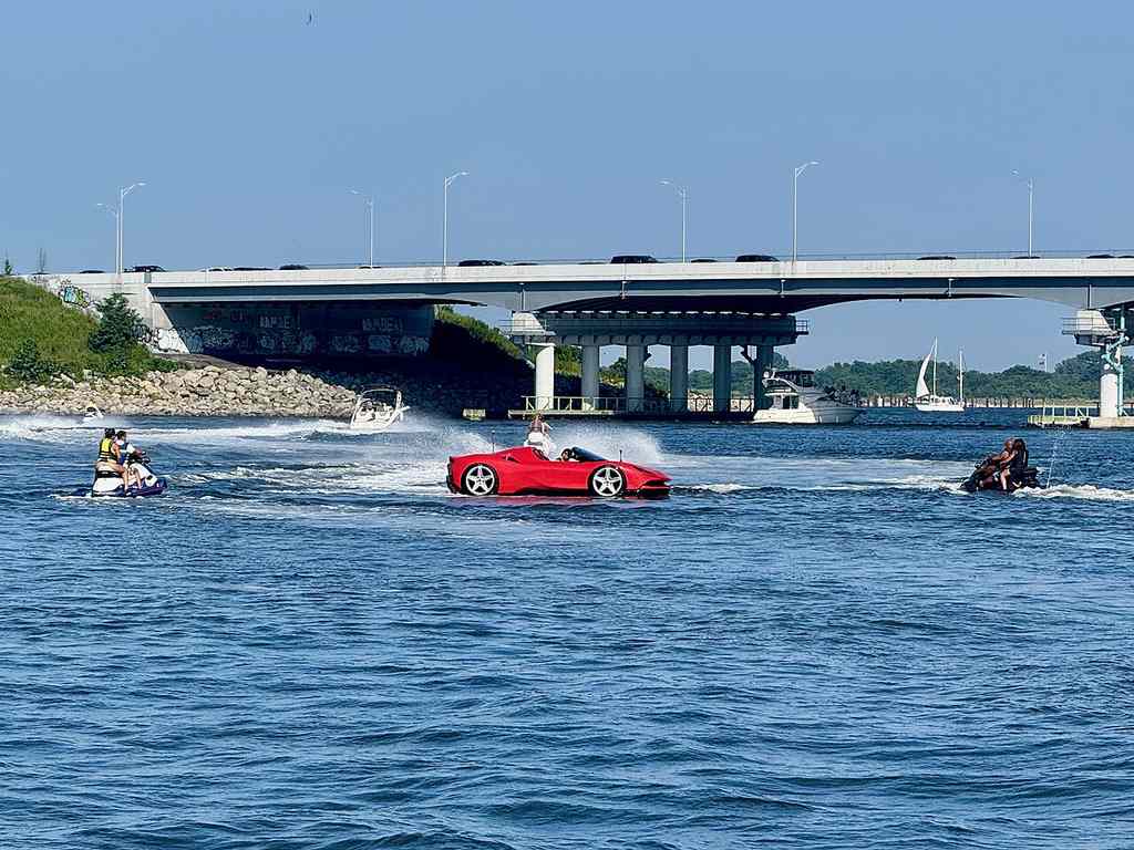 A red car-shaped boat’s speeding on the water near a bridge, with people on jet skis beside it and other boats in the background.