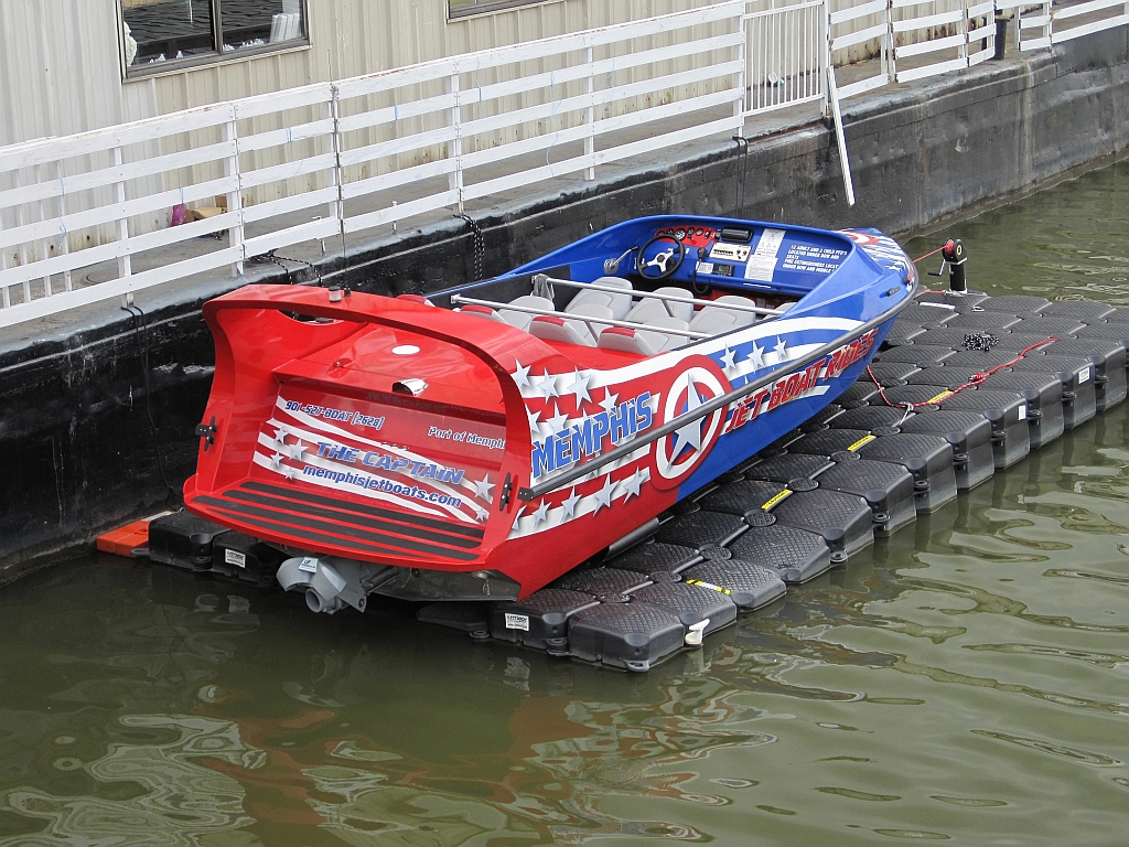 A red, white, and blue speedboat’s docked on a floating platform by a building, sporting star patterns and “Memphis” on the back.