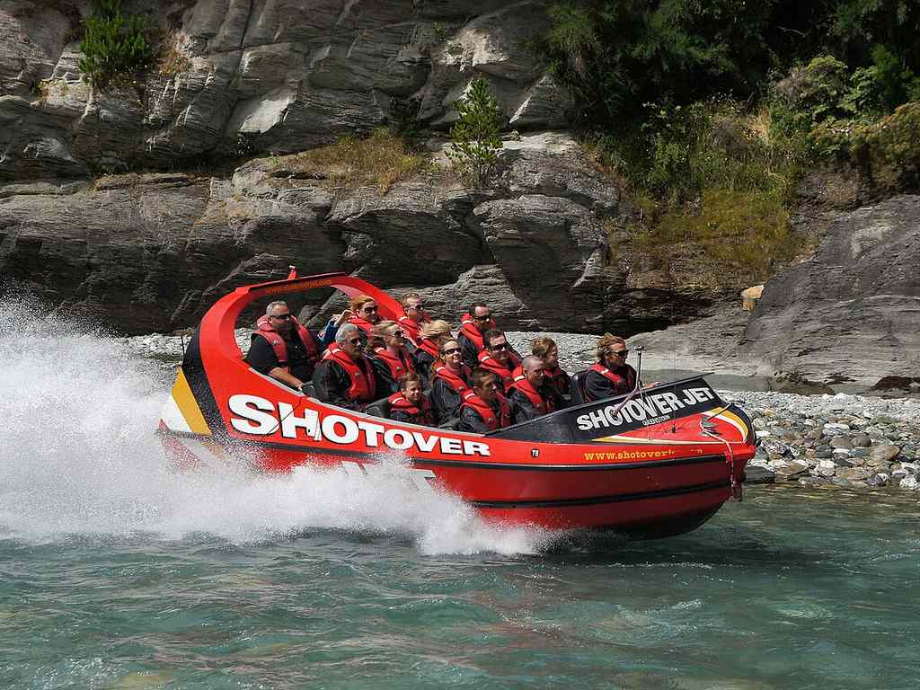 A group of people wearing life jackets ride a red jet boat labeled “Shotover” through clear water near rocky cliffs.