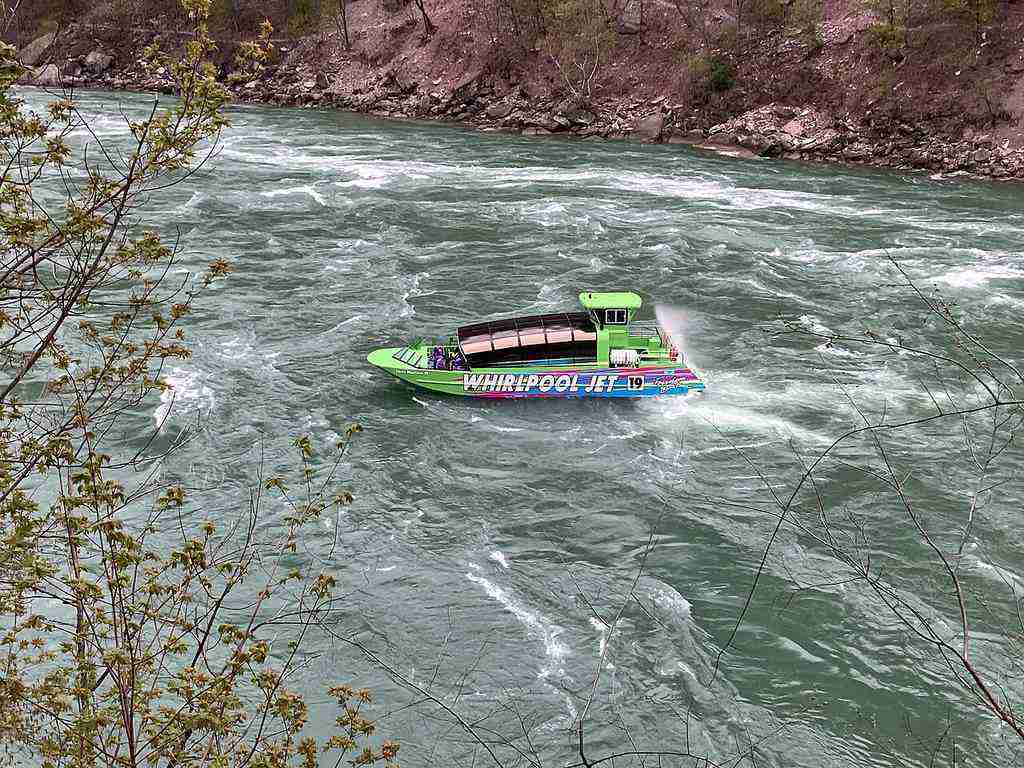 A green jet boat labeled “WHIRLPOOL JET” speeds through choppy river waters, surrounded by rocky banks lined with trees.
