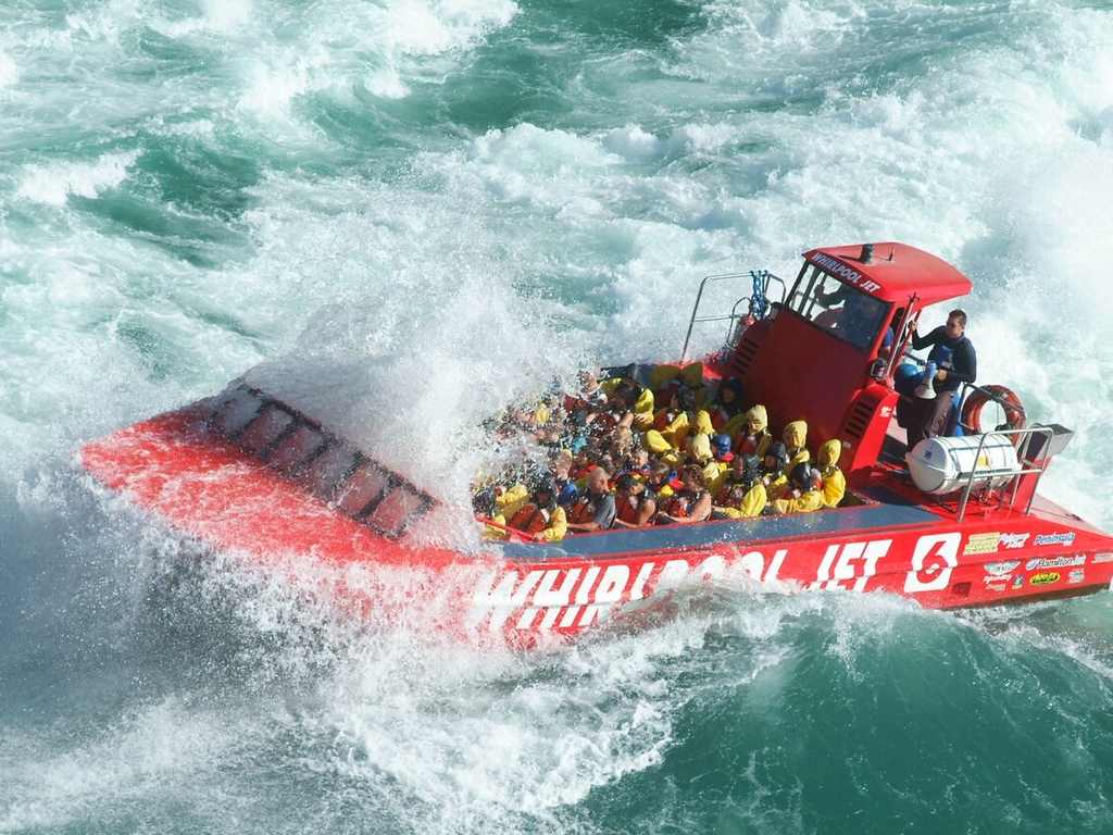 A red jet boat with passengers in yellow rain gear speeds through rough, white-water rapids, splashing water over the front of the boat.