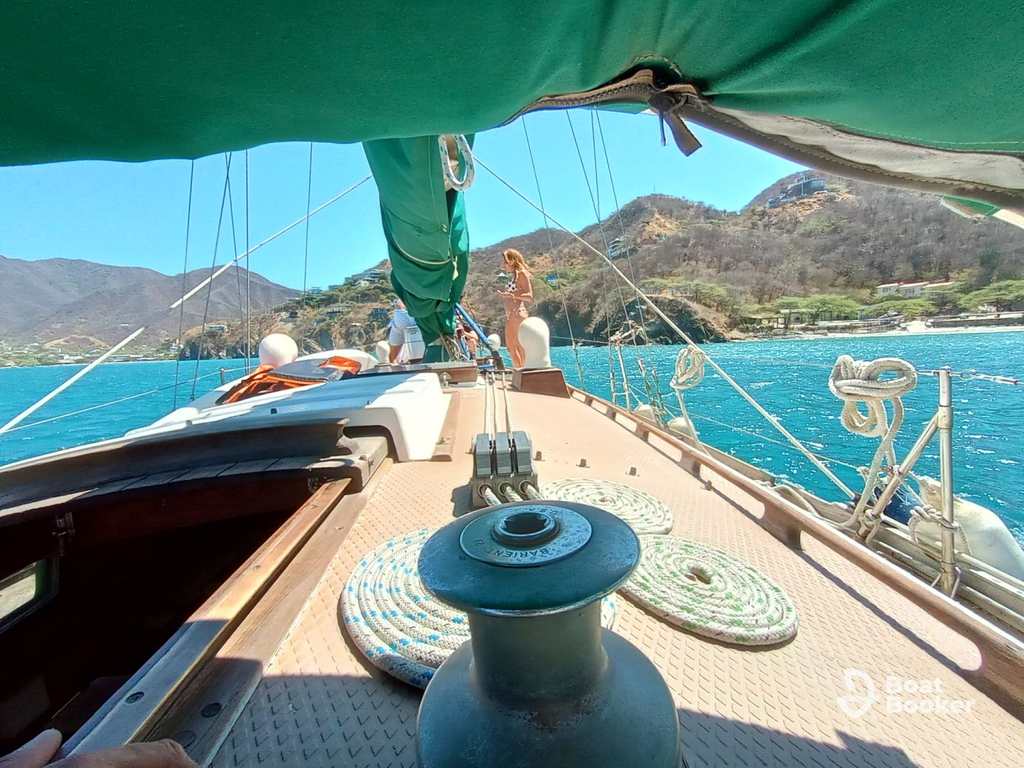 You’re on a sailboat deck heading for shore, turquoise water and mountains ahead. One person stands at the bow under a green sail, soaking up classic sailing terms in action.