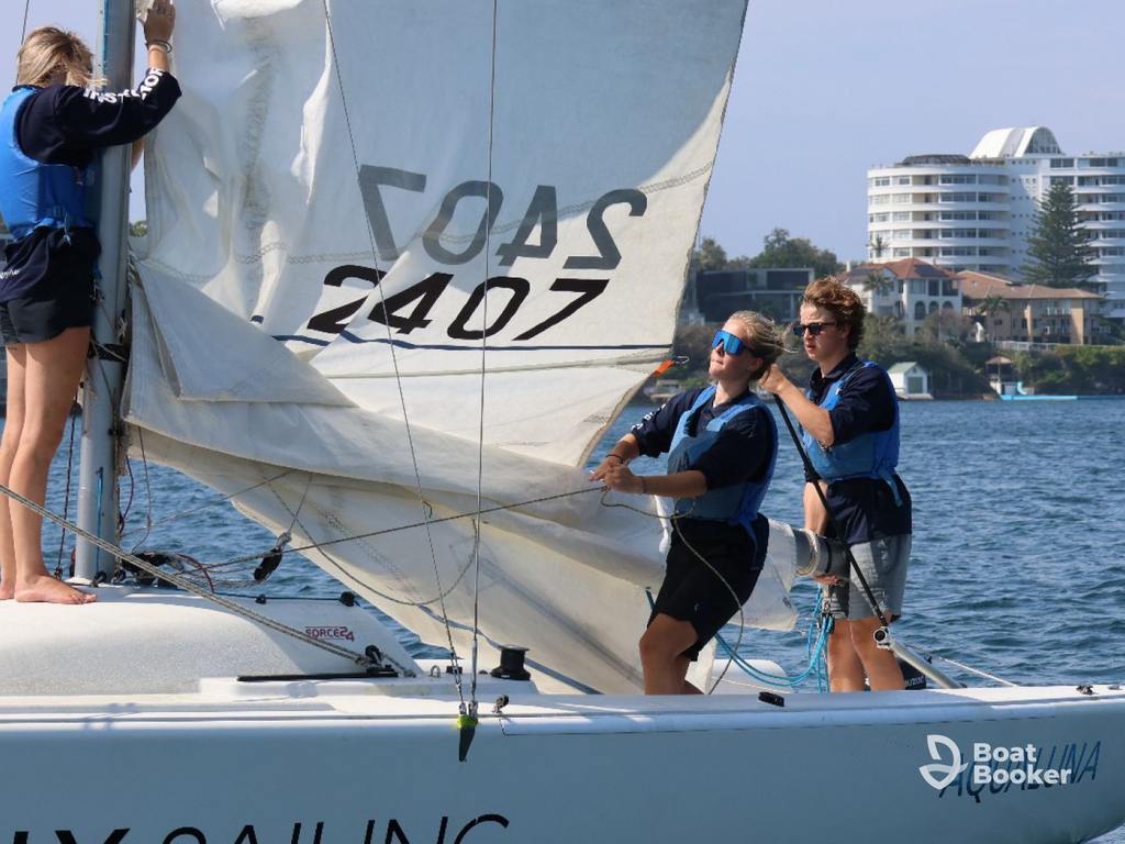 Three people are on a sailboat, adjusting the mainsail and jib on a sunny day, with water and buildings behind them. They’re showing off their sailing know-how.