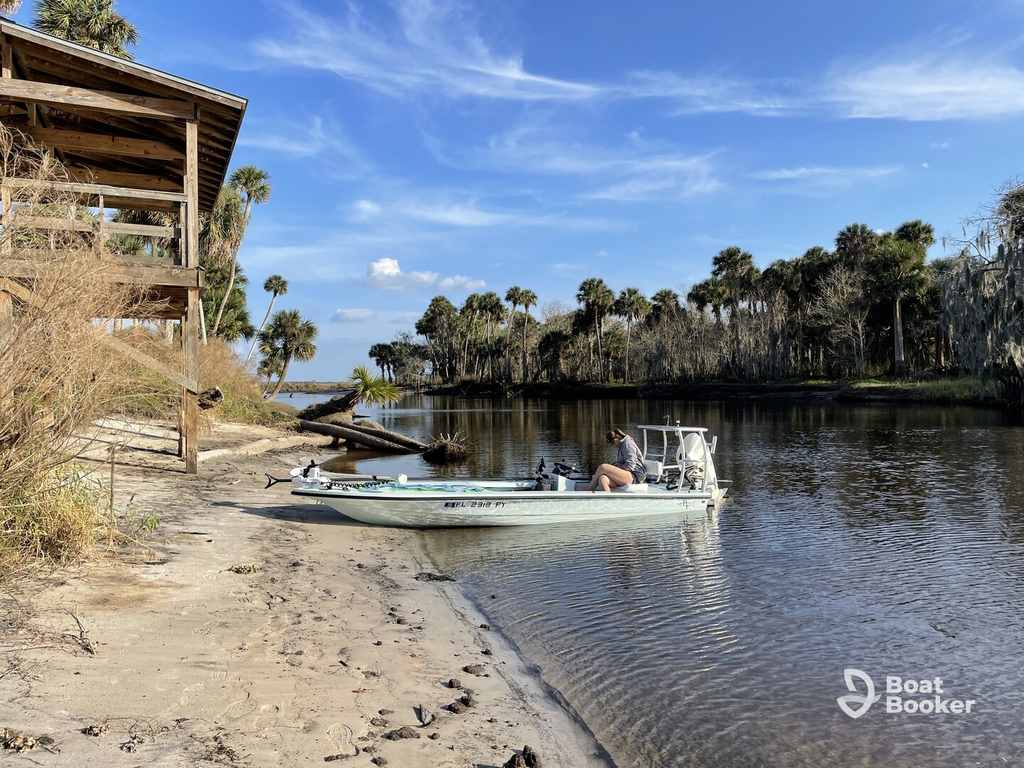 A small motorboat—great for folks with a South Carolina boating license—is docked on a sandy riverbank by a wooden structure, framed by palm trees and calm water.