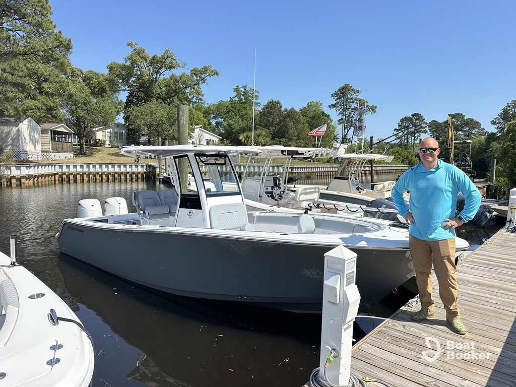 A man in a blue shirt and khaki pants stands on a dock, South Carolina boating license in hand, by a gray motorboat with twin outboards, surrounded by boats and trees.