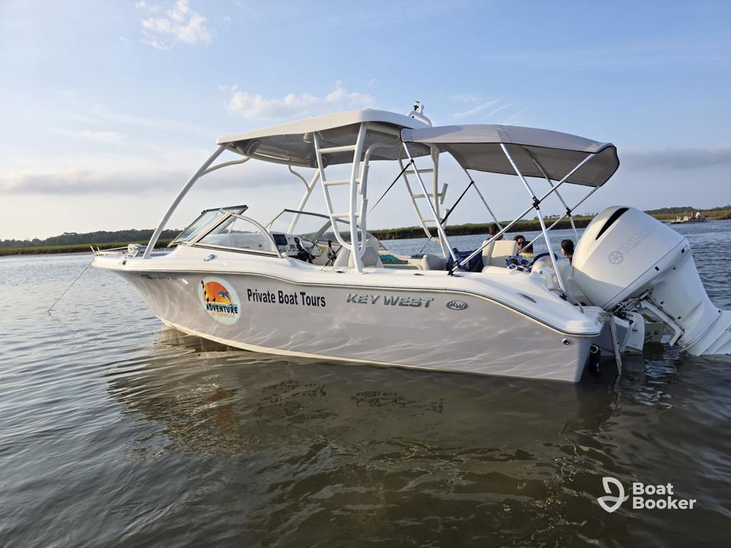A white Key West runabout called “Private Boat Tours” is anchored in shallow water by a grassy shoreline under a partly cloudy sky.