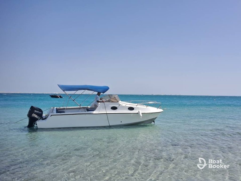 A white runabout motorboat with a blue canopy’s anchored in clear, shallow turquoise water under a clear sky. The “Boat Booker” logo’s visible in the lower right corner.