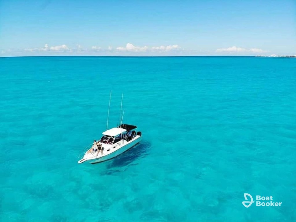 A white motorboat, like a Runabout, floats on clear turquoise water under a blue sky with distant clouds. The “Boat Booker” logo’s in the lower right corner.