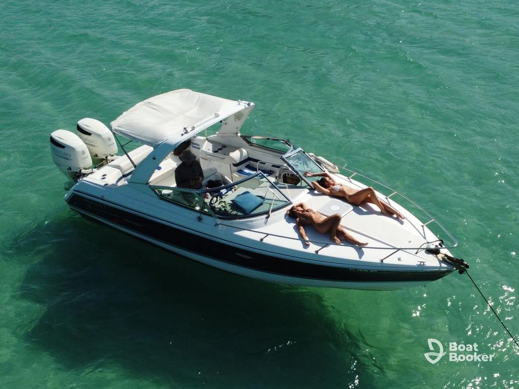 A runabout boat with a canopy floats on clear green water. Two people’re sunbathing on the bow while another’s at the helm. The Boat Booker logo’s at the bottom right.
