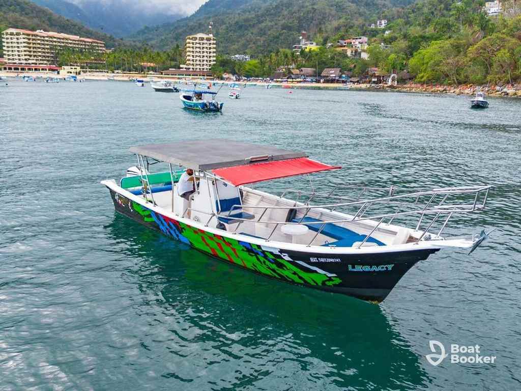 A small motorboat—maybe a Panga boat—with a canopy floats on calm water near a coastal town, green hills behind it and other boats nearby.