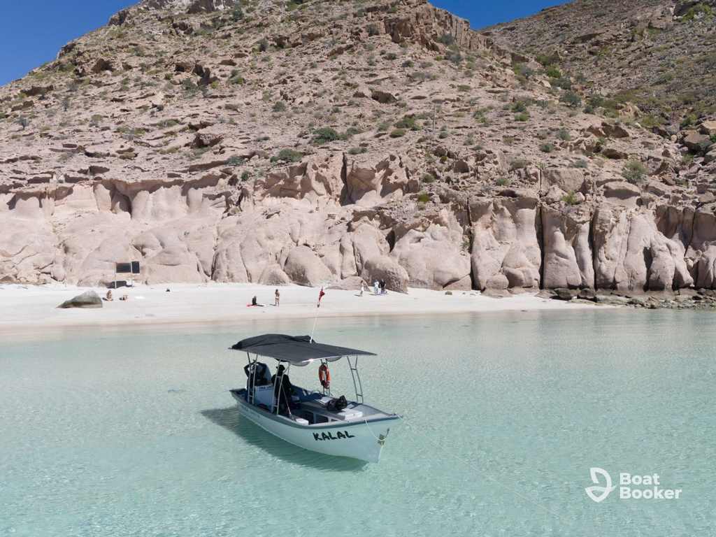 A small Panga boat floats on clear turquoise water by a rocky, sandy shore as people relax on the beach; arid hills rise beneath a blue sky.