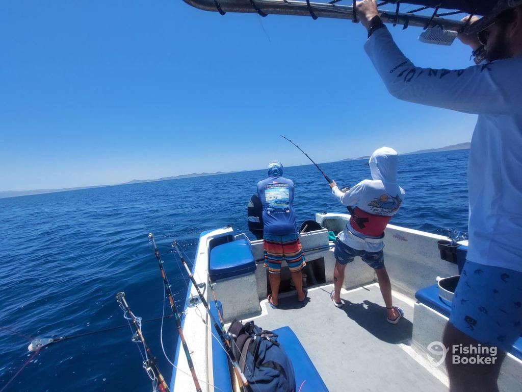 Three people are fishing on a small boat, maybe a Panga, under a clear blue sky with calm water and distant land. Two are holding fishing rods while one stands nearby.