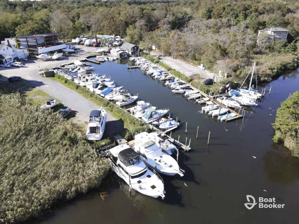 There’s a small marina with boats docked on wooden piers, surrounded by trees and parked cars—ideal if you’ve got a New York boating license. Logo at the bottom right says “Boat Booker.”