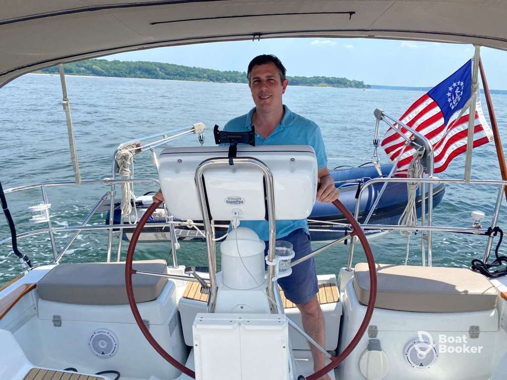Man with a New York boating license’s at the helm of a sailboat on a lake; American flag and inflatable dinghy are in the background.