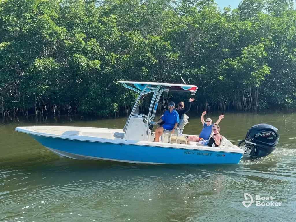 Three people ride in a small blue and white motorboat labeled “Key West,” waving as they go on calm water by dense green trees.