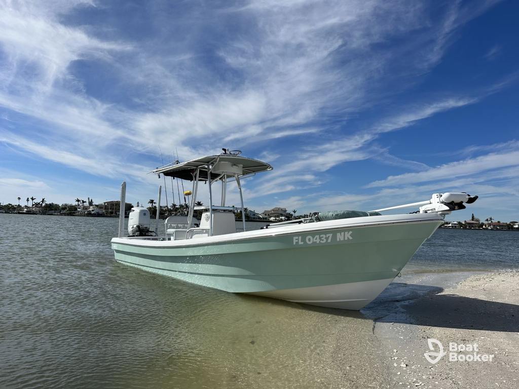 A light blue and white fishing boat with a center console’s docked on a sandy shore in shallow water, under a partly cloudy sky.