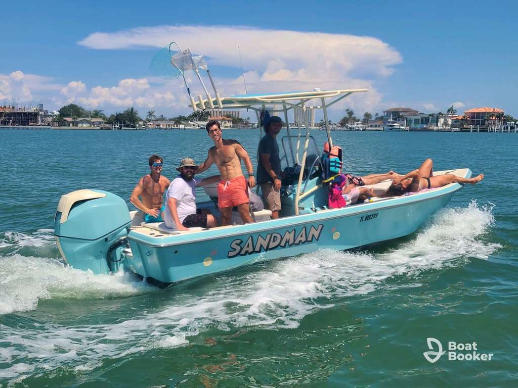 Six people are relaxin’ and smilin’ on a light blue motorboat called SANDMAN, cruisin’ on calm water by houses and trees under a partly cloudy sky.