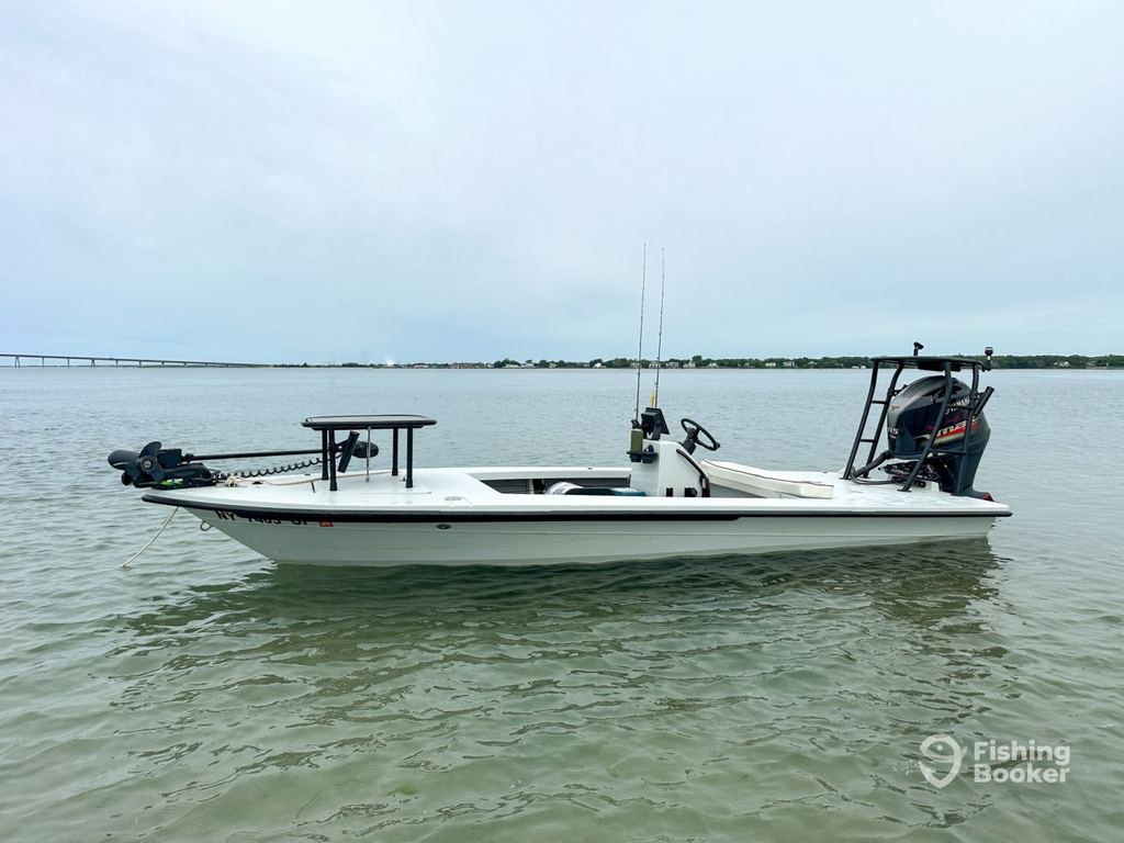 A white fishing boat with an outboard motor and rods is anchored in shallow water near shore under a cloudy sky. The “Fishing Booker” logo’s in the bottom right corner.