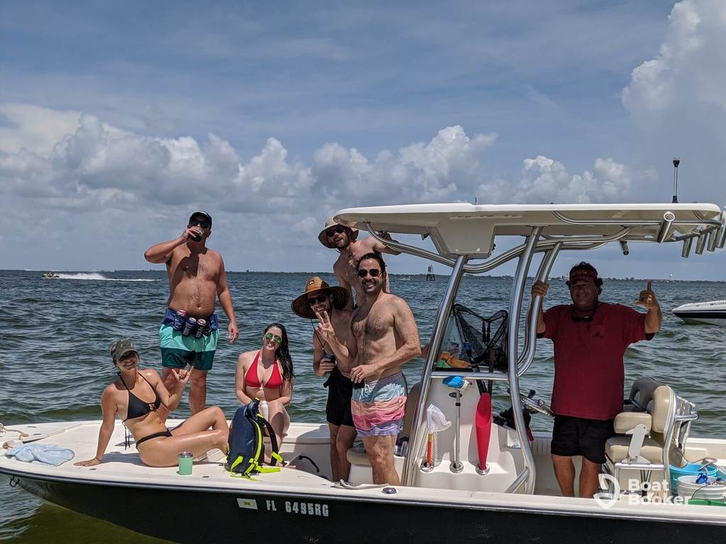 Six people in swimsuits are posing and smiling on a small motorboat in shallow water on a sunny day, with blue sky and clouds behind them.