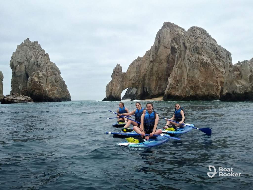 Four people in blue life vests kayak on the ocean near large, rocky cliffs beneath a cloudy sky. The water’s calm, and the cliffs rise sharply from the sea.