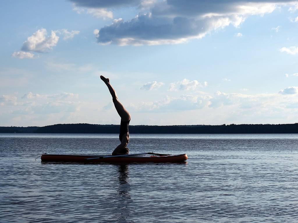 Someone’s doing a headstand on a paddleboard in calm water, with a far-off treeline and clouds showing under a blue sky.