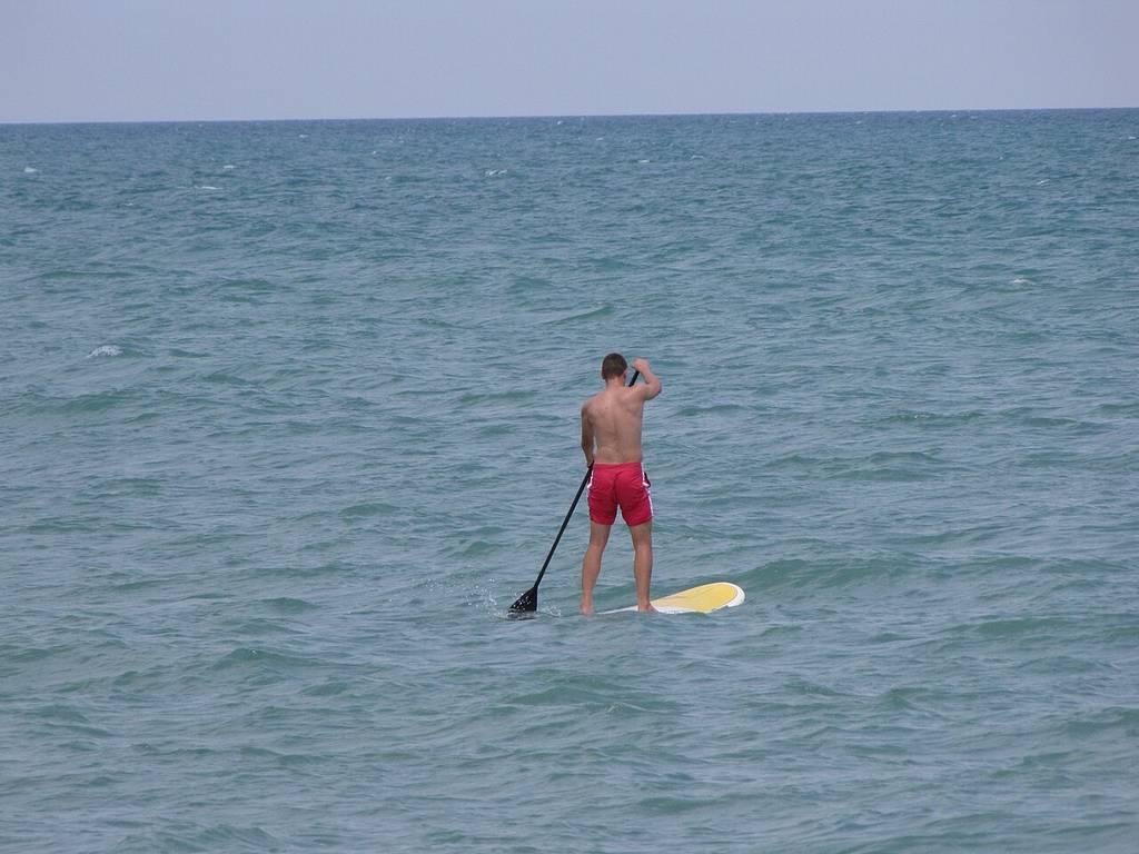 A man in red shorts paddleboards alone on calm ocean water under a clear sky.