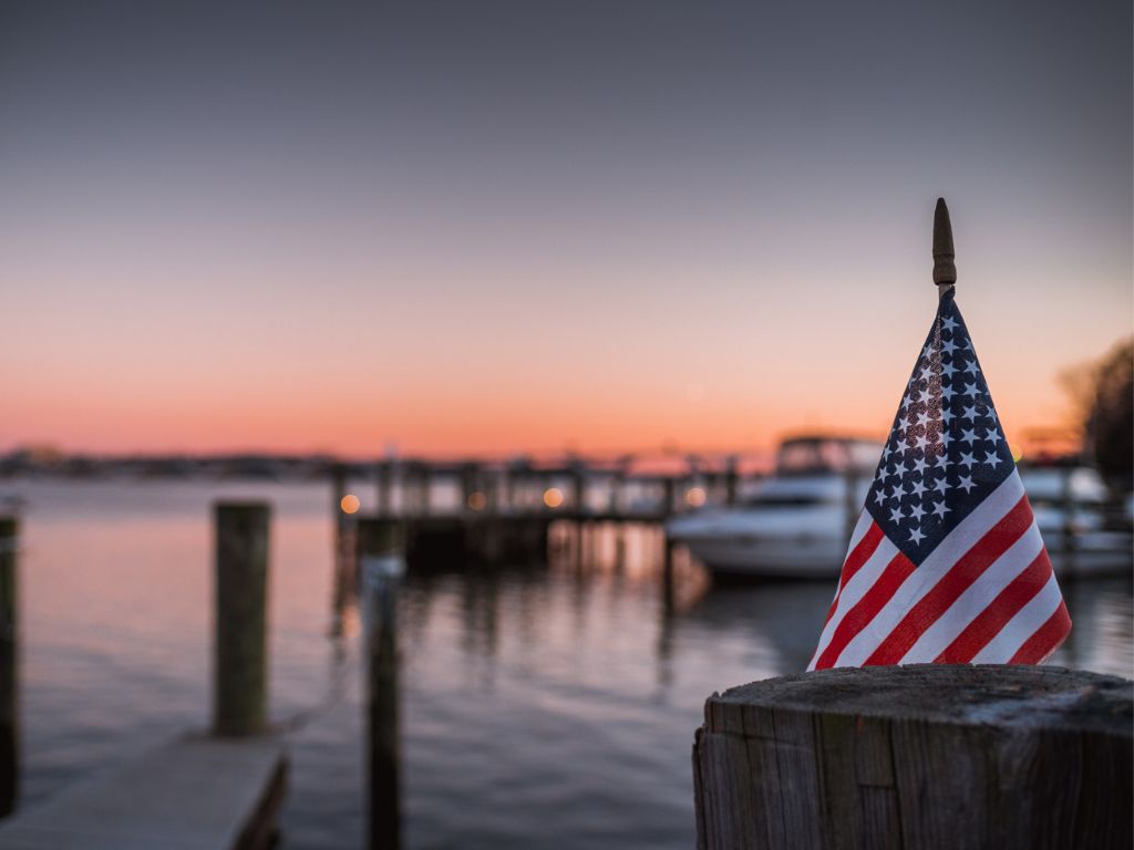 There’s a small American flag on a wooden post at a marina in one of the best boating states, with boats and docks on calm water at sunset.