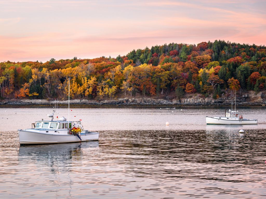 Two white fishing boats float on calm water in one of the best boating states, with an autumn-colored hill behind them under a pink and orange sky.