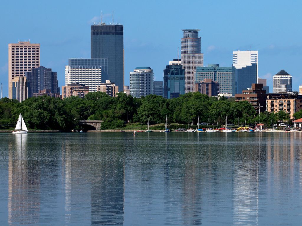You’ll see a city skyline with modern high-rises behind a lake, a sailboat, and trees up front, all under a clear blue sky—perfect for folks exploring the best boating states.