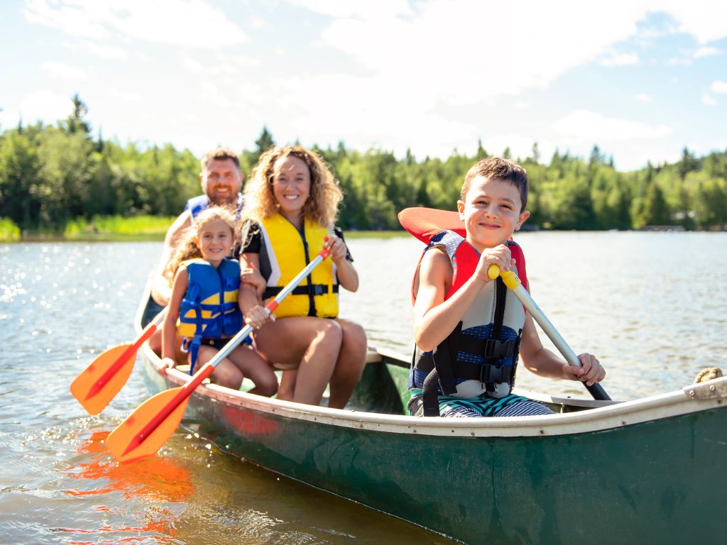 A family of four in life jackets paddles a canoe on a calm lake, surrounded by trees and blue sky. It’s one of the best boating states.