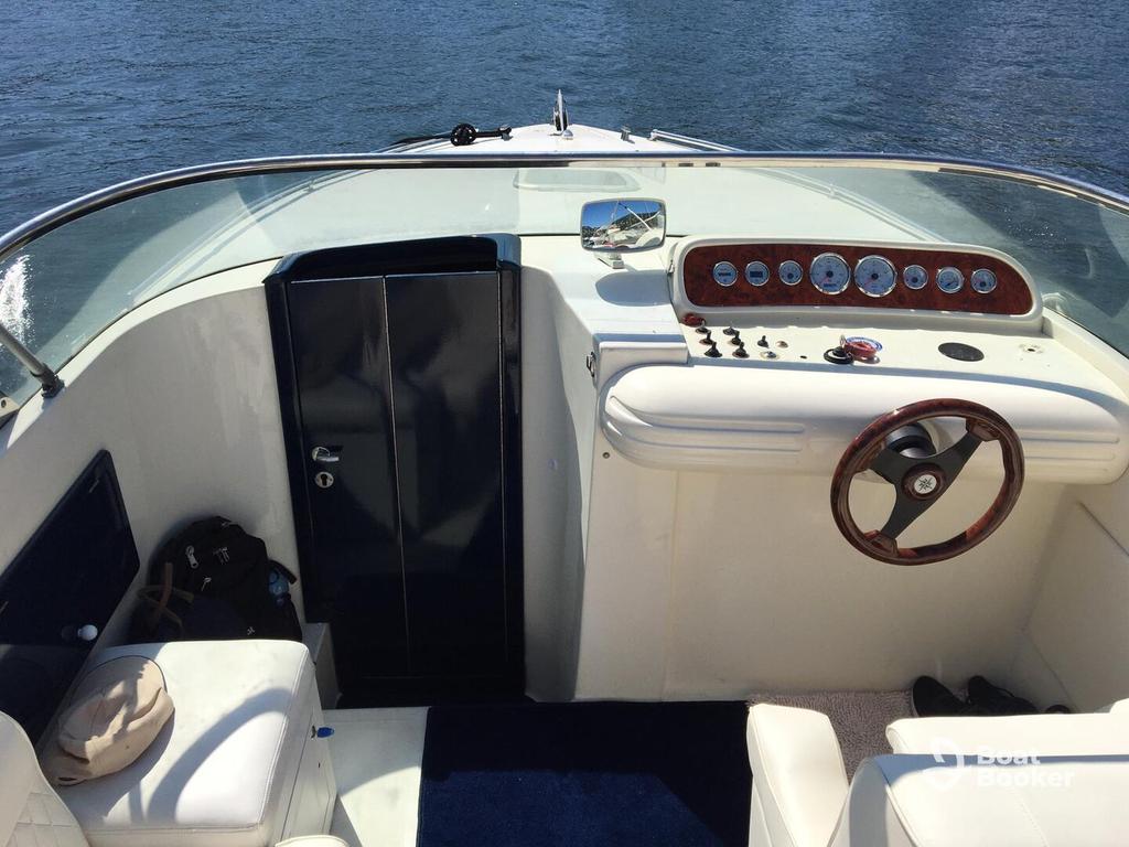 Boat cockpit with white seats, a wooden steering wheel, and dashboard gauges and controls, overlooking water on a sunny day.