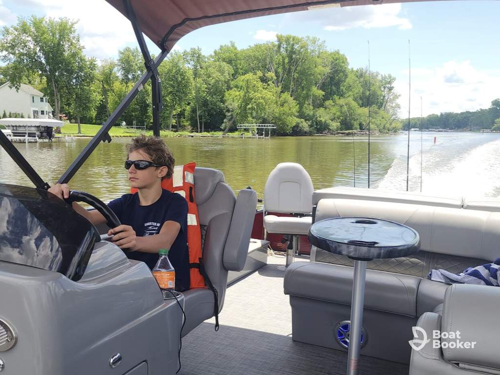 A boy with an Illinois boating license and sunglasses steers a pontoon boat on a river lined with trees, with empty seats behind him.