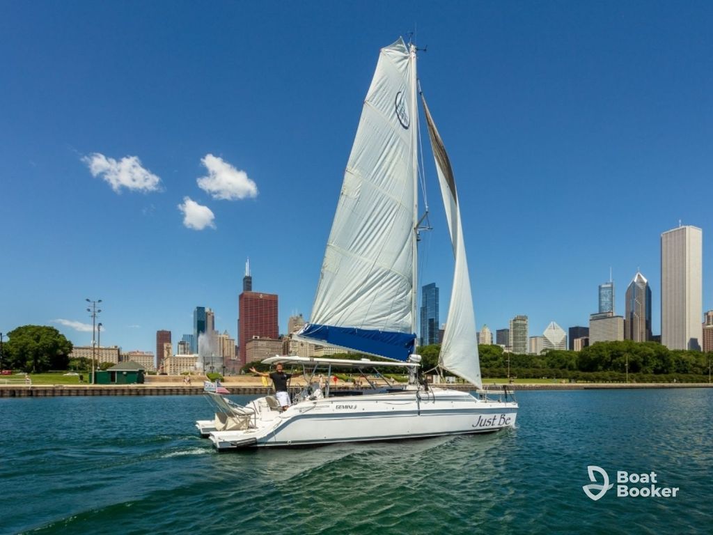 A white sailboat labeled “Just Be” sails on a lake near a city skyline, great for those with an Illinois boating license. The BoatBooker logo’s in the lower right corner.